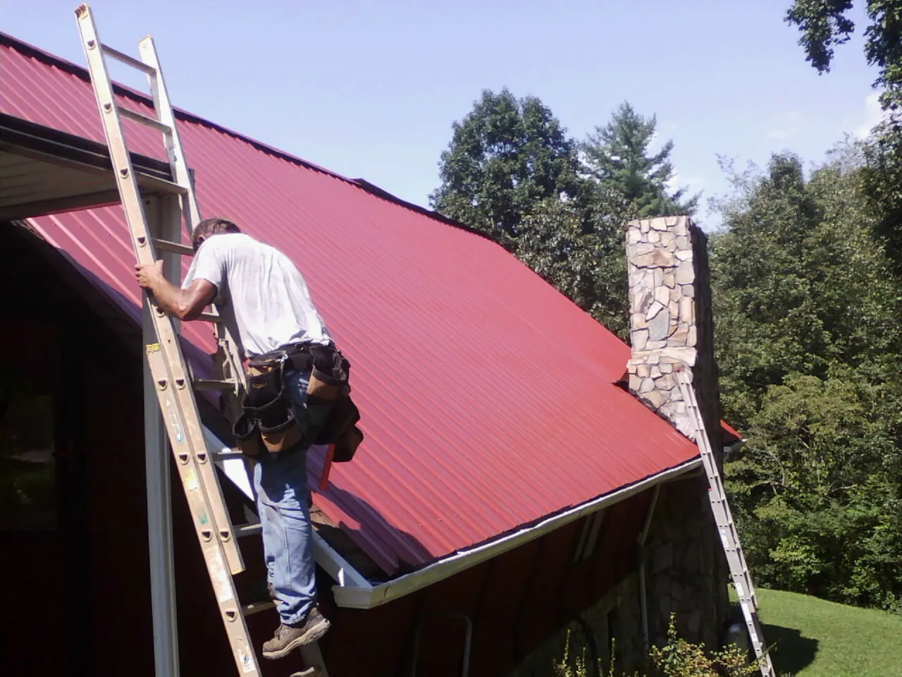 Roofer on a ladder beside a red metal roof and stone chimney — Soffit & Fascia Repair by Roofing Brothers in Thornville